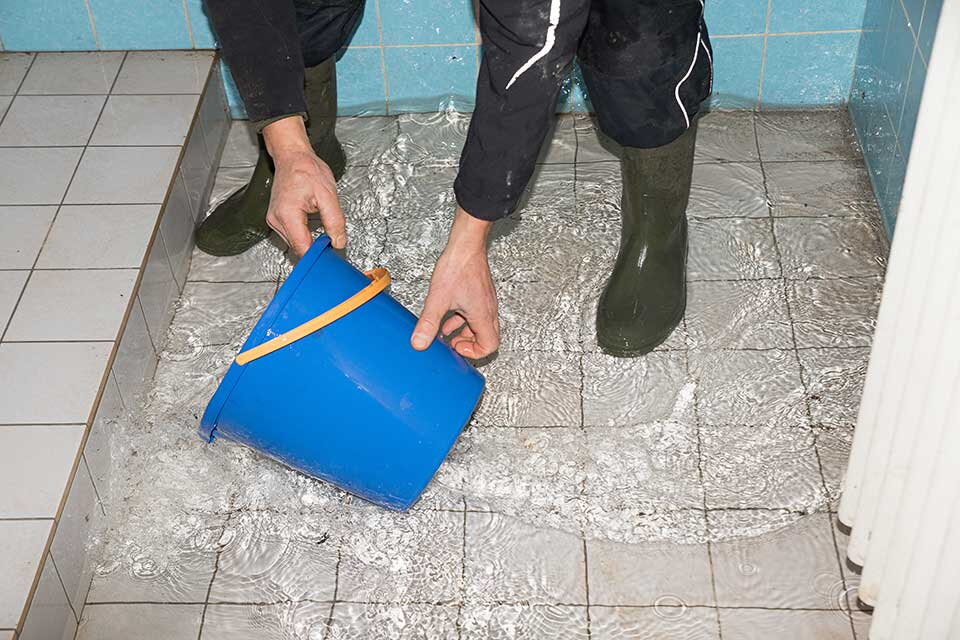 Das Bild zeigt eine Person mit Gummistiefeln, die mit einem blauen Eimer Wasser von einem gefluteten Boden aufnimmt. Der Raum hat geflieste Wände und Böden, und das Wasser steht mehrere Zentimeter hoch – es deutet auf einen Wasserschaden oder eine Übersch