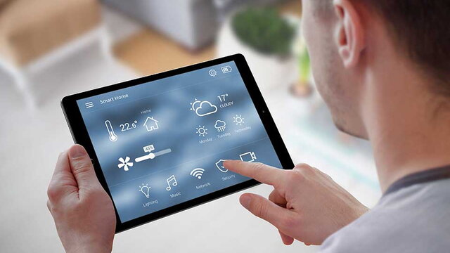 Das Bild zeigt eine Person, die ein Tablet mit einer Smart-Home-Steuerungsoberfläche in der Hand hält. Auf dem Display sind verschiedene Funktionen wie Temperaturregelung, Beleuchtung, Musik, Netzwerk, Sicherheit und Wetter angezeigt. Es symbolisiert die