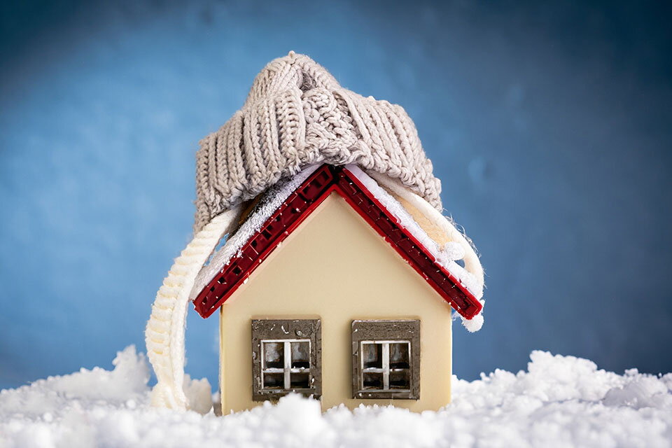 Das Bild zeigt ein kleines Modellhaus, das in Schnee steht und eine gestrickte Mütze auf dem Dach trägt. Es symbolisiert Wärmeschutz, Energieeffizienz und gute Dämmung im Winter, um das Zuhause warm und energieeffizient zu halten.