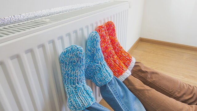 Zwei Personen liegen auf dem Boden und halten ihre Füße gegen einen weißen Heizkörper. Man sieht lediglich Beine und Füße. Ein Person trägt rote, die andere Person blaue Wollsocken.