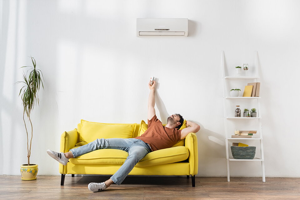 Ein junger Mann sitzt unter einem an der Wand befestigten Klimagerät auf einem gelben Sofa. Er richtet eine Fernbedienung auf das Gerät.