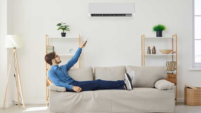 Das Bild zeigt einen Mann, der entspannt auf einem Sofa liegt und mit einer Fernbedienung eine Klimaanlage an der Wand bedient. Die Szene spielt in einem modernen, hell eingerichteten Wohnzimmer und symbolisiert Komfort und effiziente Raumklimatisierung d