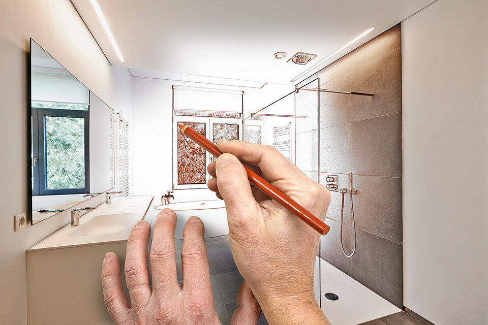 Eine Hand mit einem Stift zeichnet Planungslinien in ein Foto mit einem Badezimmer (Symbolfoto)