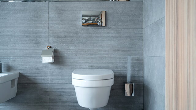 Blick auf eine grau geflieste Badezimmerwand. Man erkennt eine Toilette und verchromte Armaturen.