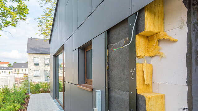 Das Bild zeigt die Fassade eines modernen Gebäudes mit sichtbarem Wandaufbau. Zu sehen ist eine mehrschichtige Konstruktion mit Dämmmaterial aus gelber Mineralwolle, schwarzer Schutzschicht und grauen Fassadenplatten. Diese Bauweise steht exemplarisch für