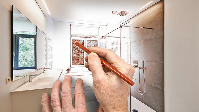 Eine Hand mit einem Stift zeichnet Planungslinien in ein Foto mit einem Badezimmer (Symbolfoto)