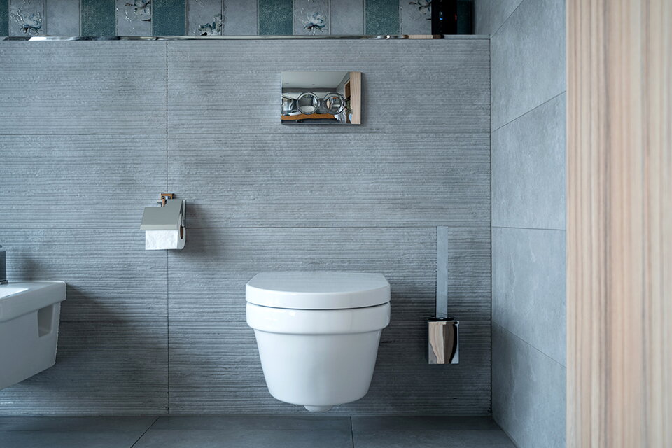 Blick auf eine grau geflieste Badezimmerwand. Man erkennt eine Toilette und verchromte Armaturen.