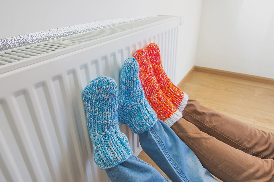 Zwei Personen liegen auf dem Boden und halten ihre Füße gegen einen weißen Heizkörper. Man sieht lediglich Beine und Füße. Ein Person trägt rote, die andere Person blaue Wollsocken.