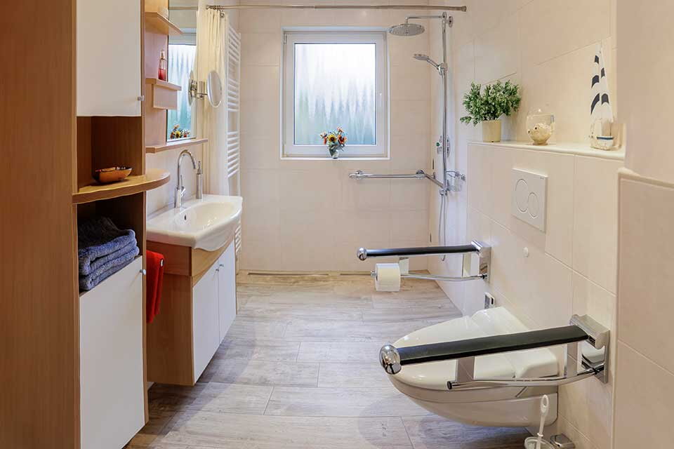 Das Bild zeigt ein barrierefreies Badezimmer mit hellen Fliesen und Holzelementen. Zu sehen sind eine bodengleiche Dusche mit Haltegriffen, ein Waschbecken mit Stauraum darunter und eine Toilette mit seitlichen Stützgriffen. Das Fenster bringt Tageslicht
