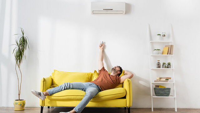 Ein junger Mann sitzt unter einem an der Wand befestigten Klimagerät auf einem gelben Sofa. Er richtet eine Fernbedienung auf das Gerät.