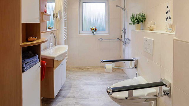 Das Bild zeigt ein barrierefreies Badezimmer mit hellen Fliesen und Holzelementen. Zu sehen sind eine bodengleiche Dusche mit Haltegriffen, ein Waschbecken mit Stauraum darunter und eine Toilette mit seitlichen Stützgriffen. Das Fenster bringt Tageslicht