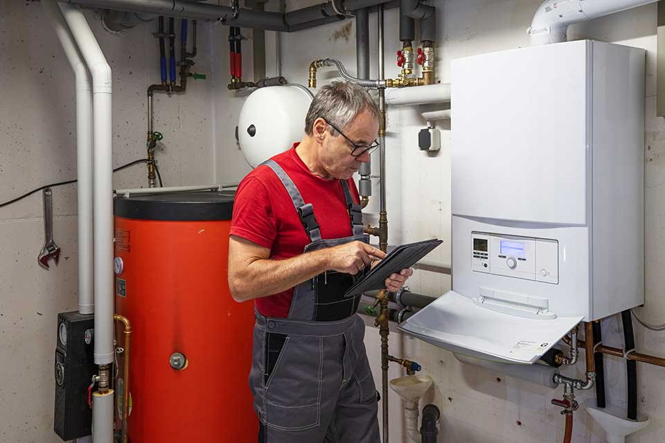 Das Bild zeigt einen Handwerker in Arbeitskleidung, der mit einem Tablet die Einstellungen oder Wartung einer Heizungsanlage überprüft. Im Hintergrund sind Rohre, ein großer roter Warmwasserspeicher und eine weiße Heizungssteuerung zu sehen.