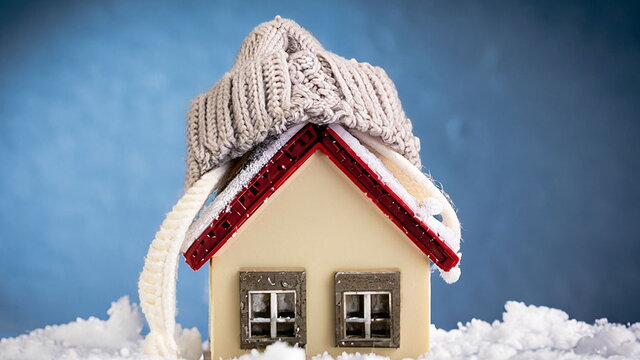 Das Bild zeigt ein kleines Modellhaus, das in Schnee steht und eine gestrickte Mütze auf dem Dach trägt. Es symbolisiert Wärmeschutz, Energieeffizienz und gute Dämmung im Winter, um das Zuhause warm und energieeffizient zu halten.